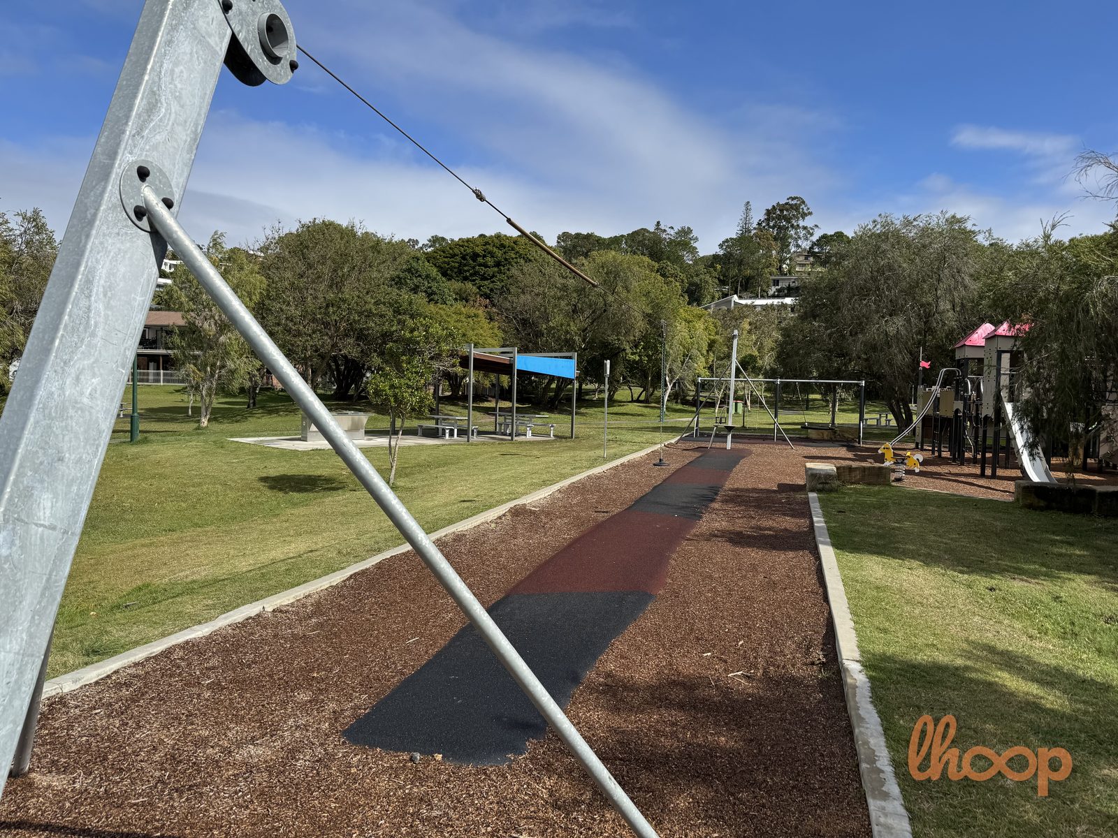 The flying fox runway on rubber softfall, its cable stretching back towards the castle, with a blue-roofed picnic shelter to the left.