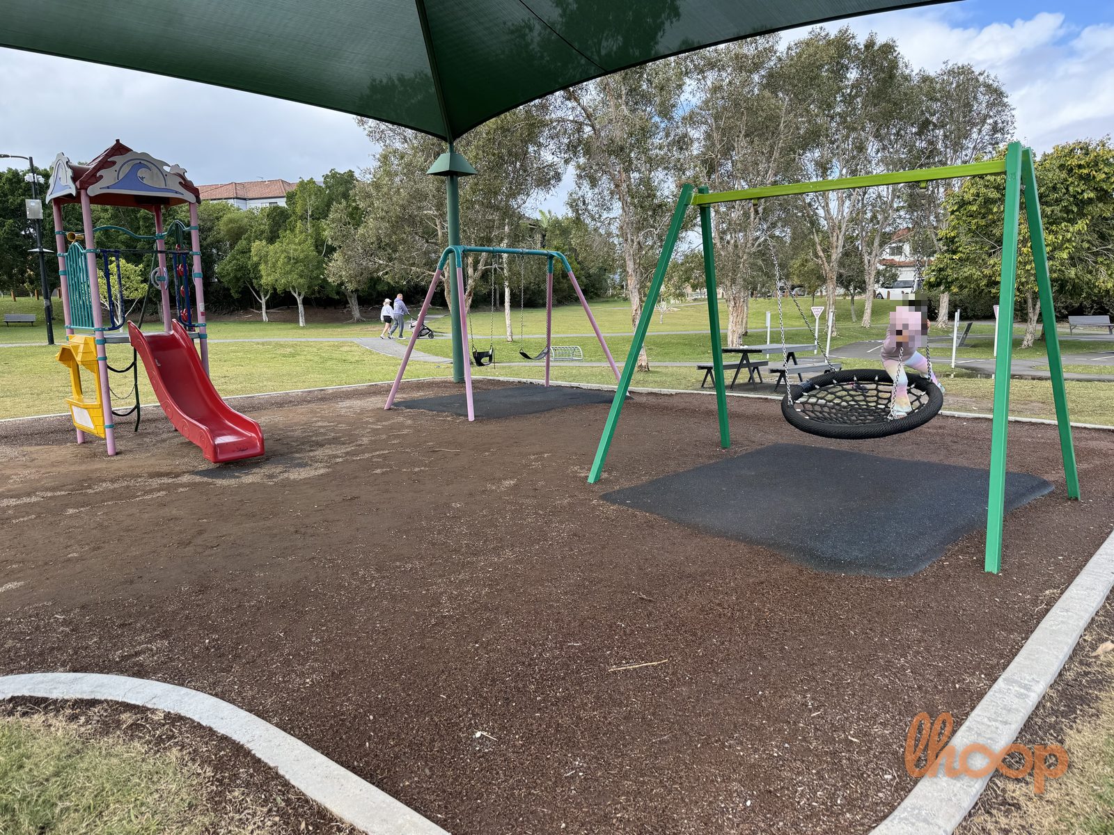The playground's red slide tower and a nest swing under the shade sail, with a picnic table and open parkland beyond.